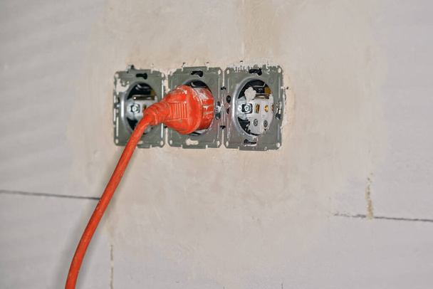 Orange power lead connected to provisional electric socket on bare wall during house construction, closeup detail. - Photo, Image