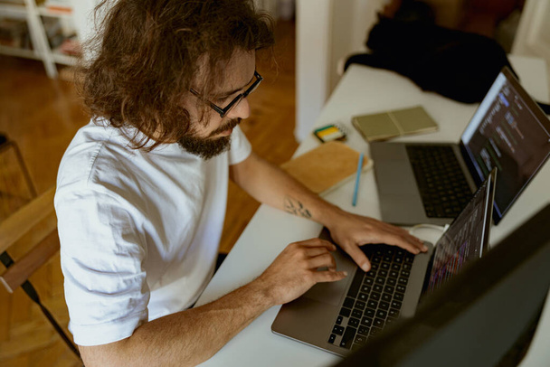 Орієнтований чоловік програміст сидить за столом і працює на ноутбуці і комп'ютері в домашньому офісі
 - Фото, зображення