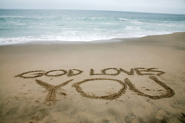Bóg kocha cię napisane w piasku plaży z oceanem jako tłem.  - Zdjęcie, obraz