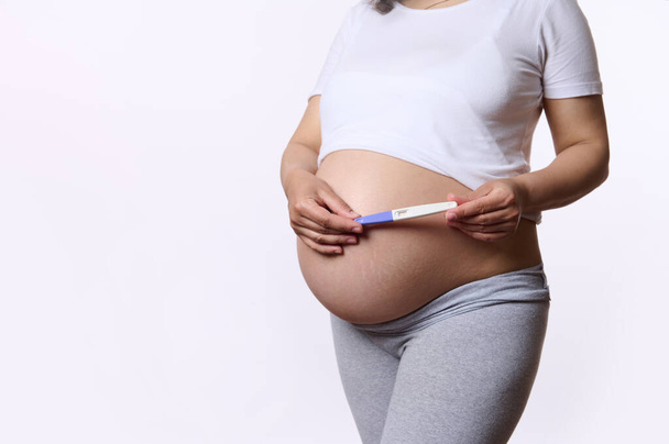 Крупный план гравийная женщина, будущая мать, держащая набор для тестов на беременность струи над ее беременным животом, изолирована на белом фоне. Плодородие. Здоровье женщин. Ожидание ребенка. Беременность. Материнство - Фото, изображение