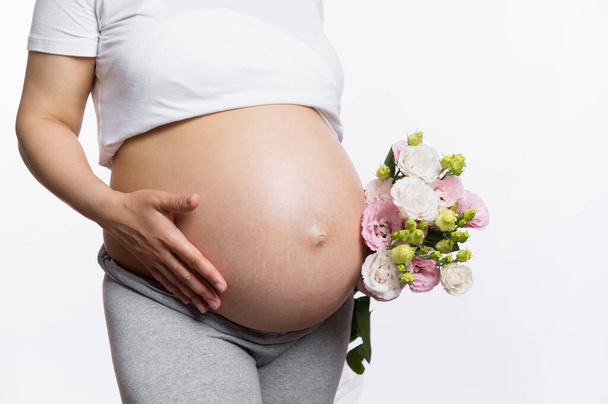 Nahaufnahme des Studios, aufgenommen von einem schwangeren Bauch einer graviden Frau mit einem Blumenstrauß, isoliert auf weißem Studiohintergrund. Gesunde Schwangerschaft, menschliche Fruchtbarkeit, Gynäkologie und Mutterschaftskonzept. Öko-Banner - Foto, Bild
