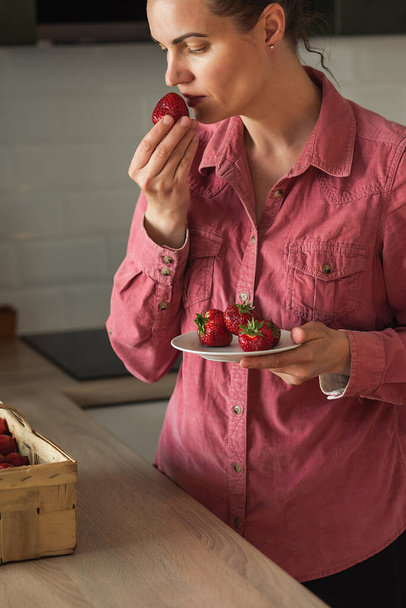 Una mujer recoge fresas de una cesta de pie en la cocina. Fresas en manos de una mujer Una mujer toma fresas de una canasta - Foto, imagen