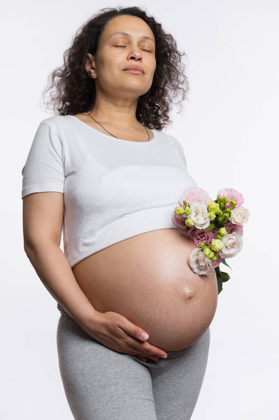 Heerlijke vrouw met een boeket lentebloemen, strelen haar grote zwangere buik, poseren met haar ogen dicht over geïsoleerde witte achtergrond, genieten van gelukkige emoties van haar zwangerschap en moederschap - Foto, afbeelding