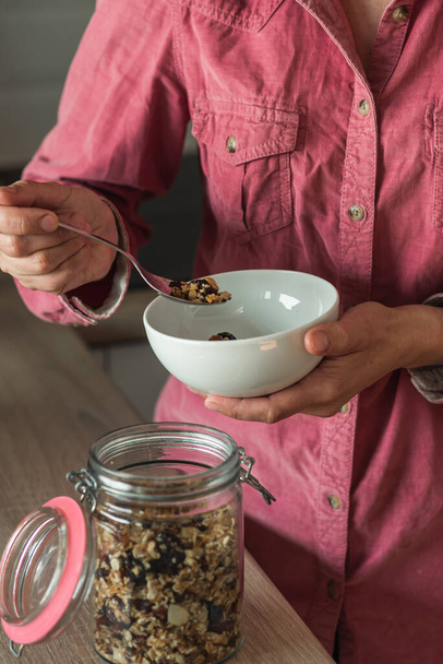 Een volwassen vrouw doet zelfgemaakte granola of muesli uit een pot in een kom. Ik maak ontbijt. Het concept van een gezond ontbijt - Foto, afbeelding