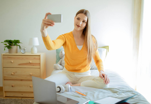 Diversión en redes sociales. Mujer joven haciendo selfie en el teléfono inteligente, mientras que el aprendizaje en línea y hacer la tarea en el ordenador portátil en el acogedor dormitorio interior. Concepto de Estudiancia Moderna - Foto, Imagen