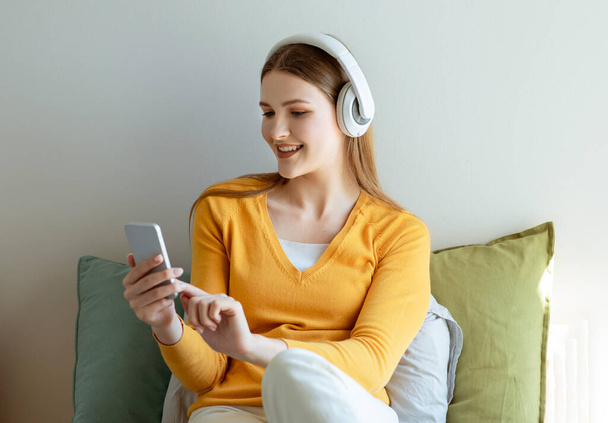 Digitaler Spaß. Junge Frau mit Kopfhörern, die mit dem Smartphone Musik hört und online in sozialen Medien im modernen Schlafzimmer im Bett sitzt. Gadgets Freizeitkonzept - Foto, Bild