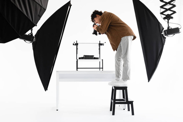 Młody człowiek content manager stojący na drabinie i robiący zdjęcia toreb pod wysokim kątem, pracujący w profesjonalnym photostudio z różnym sprzętem, widok z boku - Zdjęcie, obraz