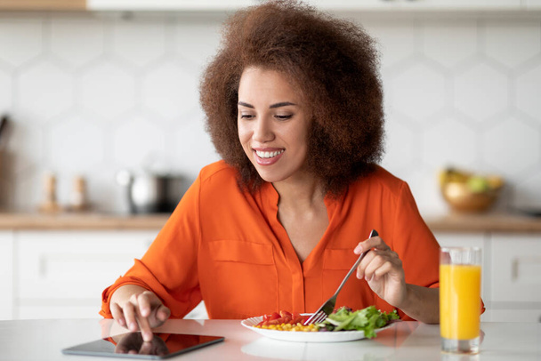 Fröhliche schwarze Frau mit digitalen Tablet-Nachrichten beim Frühstück in der Küche, lächelnde Afroamerikanerin, die am Tisch sitzt, schmackhaftes Essen genießt und im Internet surft, freier Raum - Foto, Bild