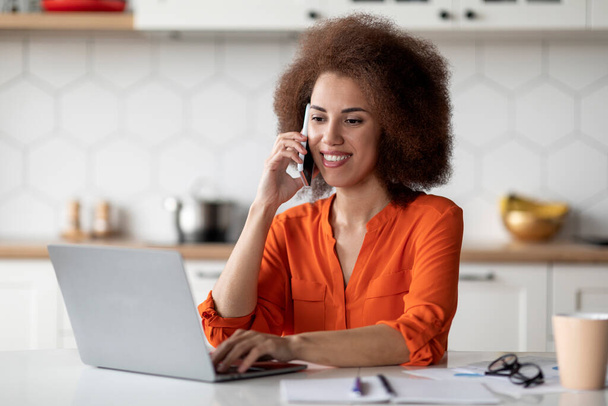 Відстань Робота. Молода чорна фрілансерка використовує мобільний телефон і ноутбук на кухні, посміхаючись афроамериканська жінка сидить за столом, працюючи з комп'ютером і маючи мобільну розмову вдома
 - Фото, зображення