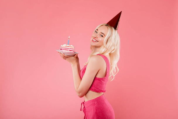 Γοητευτική χαρούμενη κυρία με καπέλο κώνου κρατώντας κομμάτι κέικ με κερί στο πάρτι γενεθλίων και χαμογελώντας στην κάμερα, ποζάροντας πάνω από ροζ φόντο στούντιο με ελεύθερο χώρο - Φωτογραφία, εικόνα