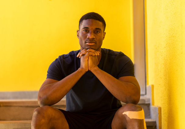 Porträt eines jungen Afroamerikaners, der auf einer Treppe sitzt und die Hände unter dem Kinn hält, mit elastischem Knieverband, und in die Kamera auf gelbem Hintergrund im Freien blickt. Horizontale Ebene. Hochwertiges Foto - Foto, Bild