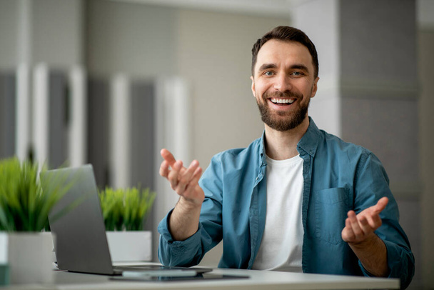 Positive Young Businessman Talking To Camera While Sitting At Desk With Laptop In Modern Office. Handsome Smiling Millennial Male Consulting Client, Enjoying Business Communication, Free Space - Fotó, kép