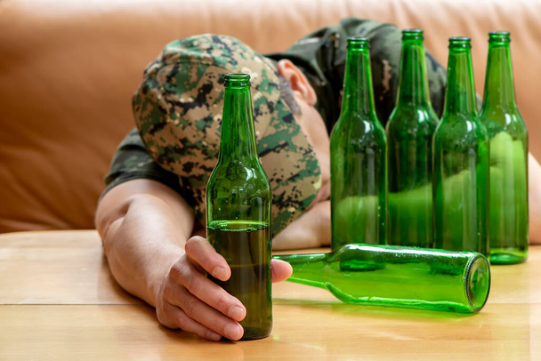 Een soldaat in uniform met een bierflesje in zijn hand viel in slaap aan tafel. Concept: oorlogsveteraan, gezinsproblemen, posttraumatisch syndroom, alcoholisme. - Foto, afbeelding