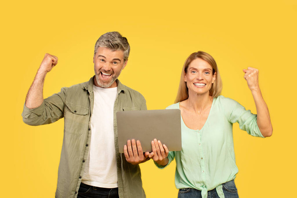 Emotional middle aged spouses using laptop on yellow studio background, raising fists up and exclaiming, celebrating success. Online bet, trading, cashback, giveaway - Photo, image