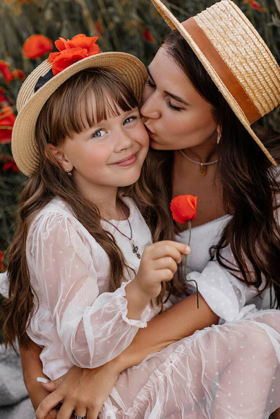 Beautiful young woman with girl in field with poppies, mother and daughter in white dresses and straw hats in evening at sunset, Summer countryside nature flowers, family relaxing, Rural simple life - Photo, Image