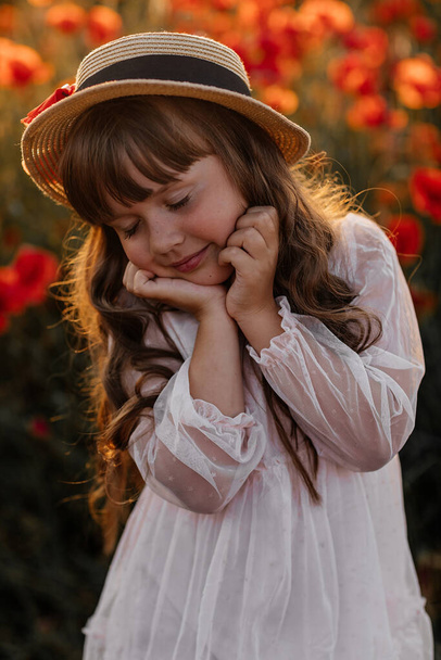 Portrait of cute girl in white dress and straw hat in field with poppies in evening at sunset, happy child, Stylish kid, Summer countryside nature flowers, Female relaxing, Rural simple life, family  - Photo, Image