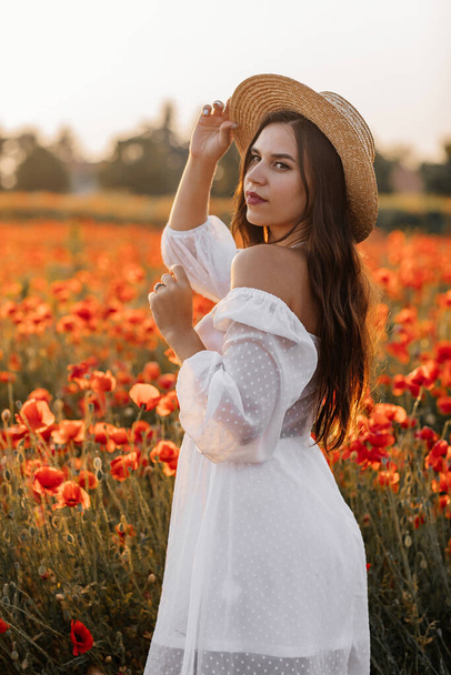 Belle jeune femme en robe blanche courte et chapeau de paille dans le champ avec des coquelicots le soir au coucher du soleil et tient le coquelicot à la main, Summer campagne nature fleurs, Femme détente, Rural vie simple - Photo, image