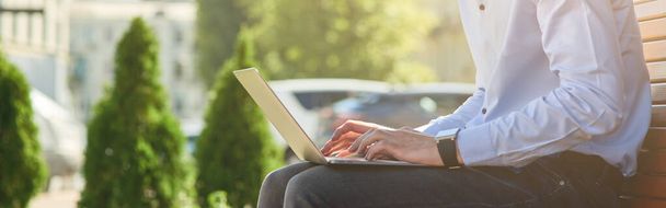 Vue latérale d'un beau mâle freelance ou homme d'affaires travaillant en ligne sur ordinateur portable tout en étant assis sur le banc à l'extérieur par une journée ensoleillée. Concept d'emploi à distance - Photo, image