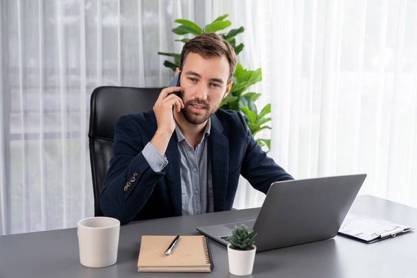 Fleißiger und fleißiger Geschäftsmann, der an seinem Laptop arbeitet, während er mit Kunden auf seinem Mobiltelefon spricht und den modernen Multitasking-Büroarbeiterlebensstil präsentiert. Einheit - Foto, Bild