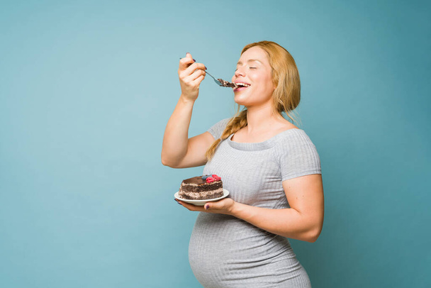 Attractive pregnant woman eating a big slice of cake with her eyes closed in a studio - Fotoğraf, Görsel