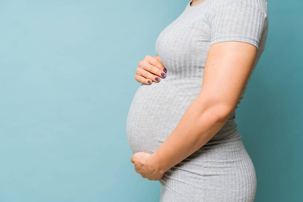Profile view of the body of a pregnant woman embracing her belly in a studio with a blue background - Photo, Image