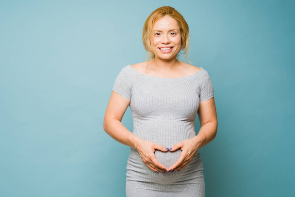 Mulher grávida caucasiana atraente olhando feliz ao fazer uma forma de coração em sua barriga em um fundo azul - Foto, Imagem