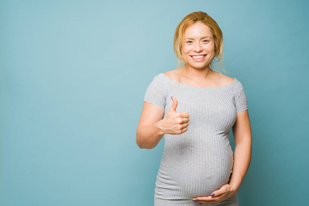 Όμορφη έγκυος γυναίκα στα 30 της δίνει τους αντίχειρες της ως ένα τραγούδι της έγκρισης, ενώ στέκεται πάνω σε ένα μπλε φόντο - Φωτογραφία, εικόνα