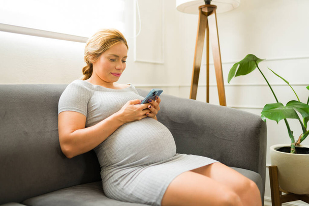 Приваблива жінка використовує мобільний додаток для відстеження її прогресу під час вагітності, сидячи у своїй вітальні
 - Фото, зображення