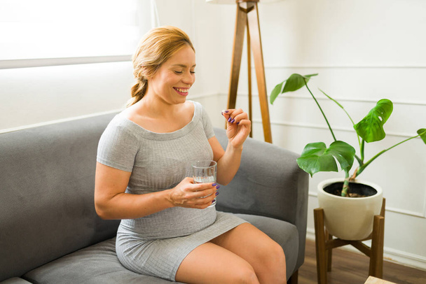 Портрет красивой и веселой беременной женщины, принимающей добавки к беременности и улыбающейся в гостиной - Фото, изображение