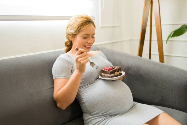 Attraktive Frau genießt ihre Schwangerschaft und isst als eine ihrer Gelüste ein großes und köstliches Stück Kuchen - Foto, Bild