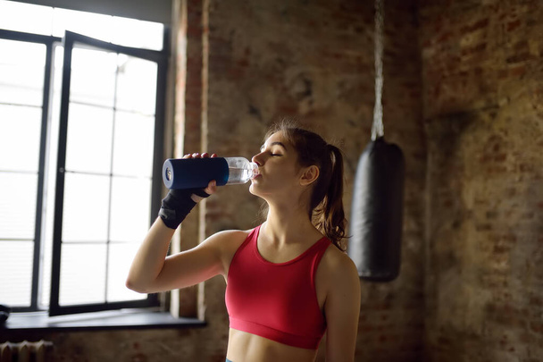 Portrait of young beautiful athlete in a boxing gym. A boxer girl drinking beverage from a bottle during a workout. In the process of training need to drink a lot water. Healthy lifestyle - Foto, imagen