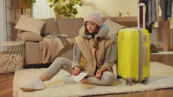 Egy csalódott fiatal nő kalapban, bőrönddel a szőnyegen ülve. Egy szomorú nő utazó boldogtalan egy sikertelen utazással. Utazási bürokrácia. Útlevélproblémák. - Felvétel, videó