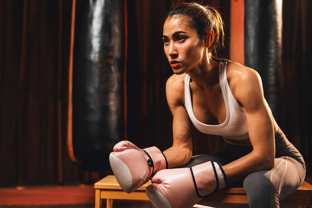 Ασιάτισσα γυναίκα Muay Thai πυγμάχος ή kickboxing λαμβάνοντας σύντομο διάλειμμα κάθεται με τα γάντια της στο γυμναστήριο με τον εξοπλισμό πυγμαχίας στο παρασκήνιο. Δυνατή και μυώδης αθλήτρια του σώματος. Ιμπέτους - Φωτογραφία, εικόνα
