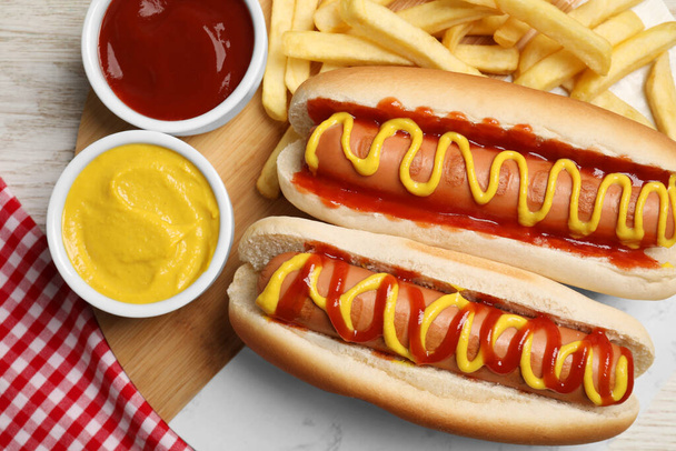 Delicious hot dogs with mustard, ketchup and potato fries on white wooden table, flat lay - 写真・画像