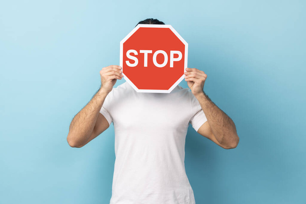 Portrait of unknown man wearing white T-shirt covering face with Stop symbol, anonymous person holding red traffic sign, warning about road safety rules. Indoor studio shot isolated on blue background - Fotoğraf, Görsel