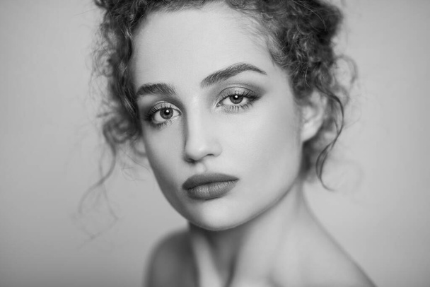 Black and white beauty portrait of young confident woman fashion model with collected dark curly hair and makeup, looking at camera with calm face. Indoor studio shot isolated on gray background. - Fotoğraf, Görsel
