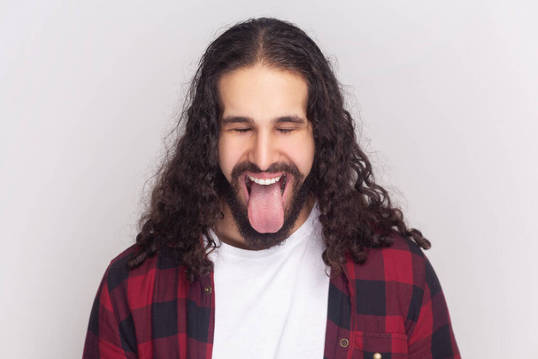 Comic bearded man in checkered red shirt sticks out tongue, makes happy grimace, expresses positive emotions, foolishes with friends. Indoor studio shot isolated on gray background. - Photo, image