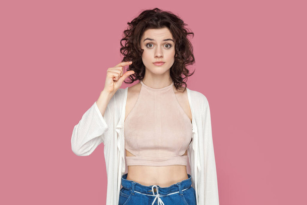 Portrait of confused disappointed beautiful woman with curly hairstyle wearing casual style outfit showing small size with fingers. Indoor studio shot isolated on pink background. - Φωτογραφία, εικόνα
