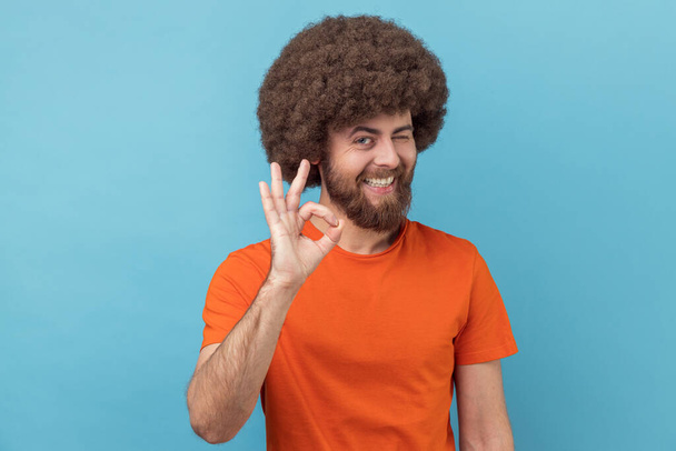 Portrait of positive man with Afro hairstyle wearing orange T-shirt standing and looking at camera with Ok sign, winking and toothy smile. Indoor studio shot isolated on blue background. - Φωτογραφία, εικόνα