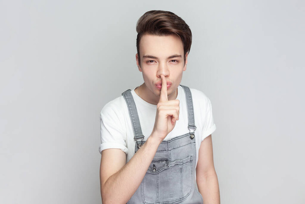Portrait of mysterious brunette man standing presses index finger to lips, makes hush gesture tells secret, asks to be quiet, wearing denim overalls. Indoor studio shot isolated on gray background. - Фото, зображення