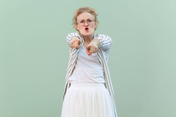 Portrait of angry shocked young adult blonde woman wearing striped shirt and skirt, pointing at you, choosing you, screaming. Indoor studio shot isolated on light green background. - 写真・画像