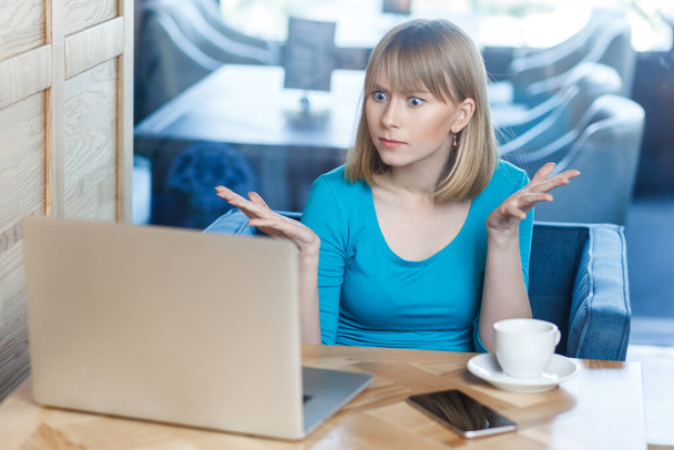 Portrait of unhappy serious attractive young woman with blonde hair in blue shirt working on laptop, having video call, spreads hands, asking how could you. Indoor shot, cafe background. - Foto, Imagen