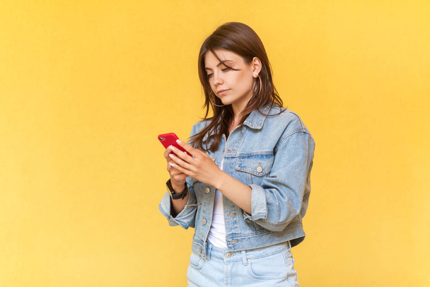 Side view portrait of serious concentrated woman wearing denim jacket using cell phone, chatting with somebody with attentive expression. Indoor studio shot isolated on yellow background. - Foto, Bild