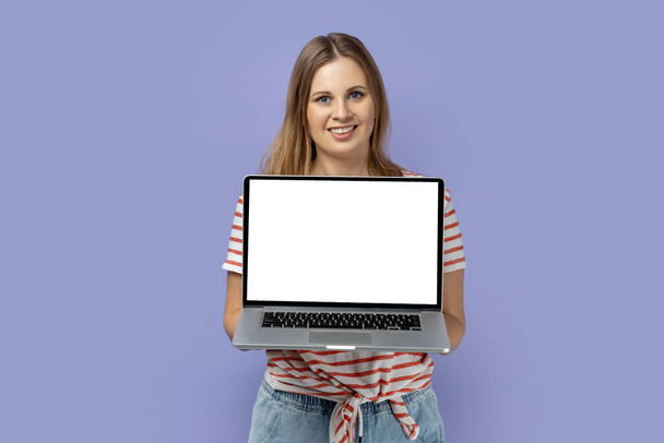 Portrait of happy joyful blond woman wearing striped T-shirt holding laptop with white empty display with empty space for advertisement. Indoor studio shot isolated on purple background. - Fotoğraf, Görsel