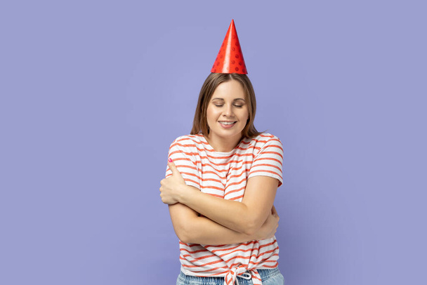 Portrait of self-satisfied egoistic woman in party cone celebrating her birthday, embracing herself and smiling with pleasure, feeling self-pride. Indoor studio shot isolated on purple background. - Foto, Bild