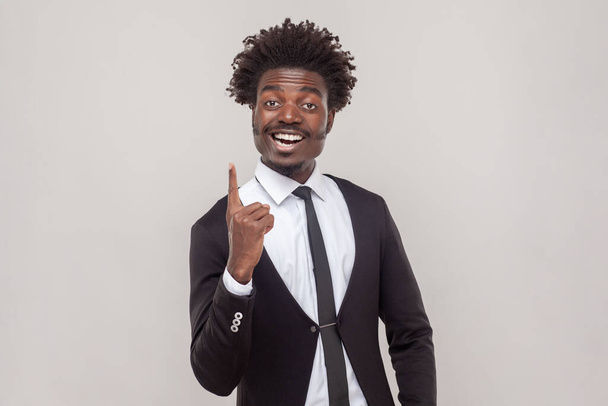 Clever man with Afro hairstyle get new idea, raises fore finger, ready to start work on project, confident in success, wearing white shirt and tuxedo. Indoor studio shot isolated on gray background. - Foto, afbeelding