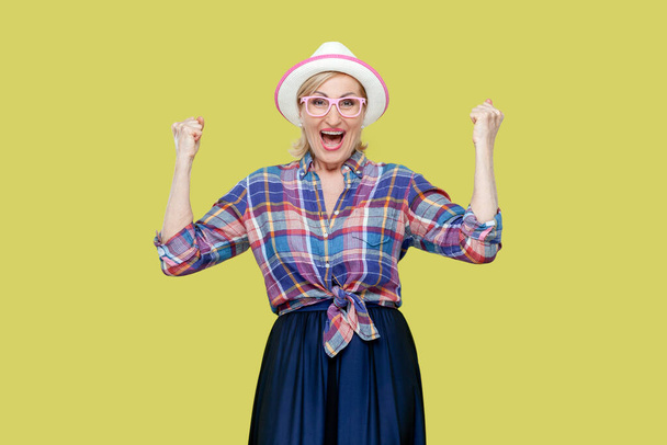 Overjoyed senior woman in checkered shirt, hat, eyeglasses clenches fists with happiness, opens mouth widely as shouts loudly, celebrates her success. Indoor studio shot isolated on yellow background. - Fotografie, Obrázek