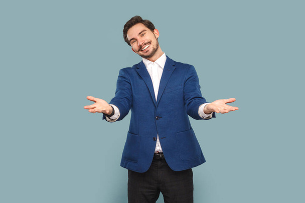 Portrait of friendly positive man with mustache standing with spread hands, saying welcome, take for free, wearing white shirt and jacket. Indoor studio shot isolated on light blue background. - Photo, image