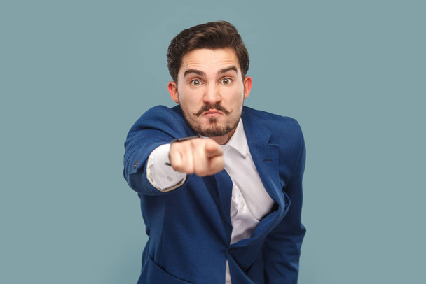 Portrait of serious man with mustache with negative facial expression pointing finger to camera, selecting you, wearing official style suit. Indoor studio shot isolated on light blue background. - Foto, Bild