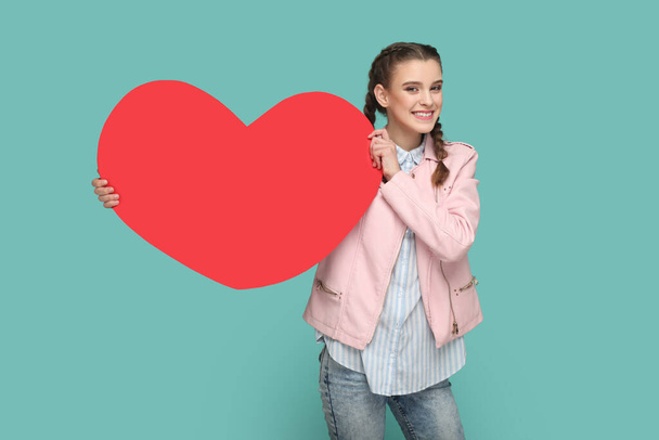 Portrait of extremely joyful cheerful teenager girl with braids wearing pink jacket, showing big red heart, smiling to camera. Indoor studio shot isolated on green background. - 写真・画像